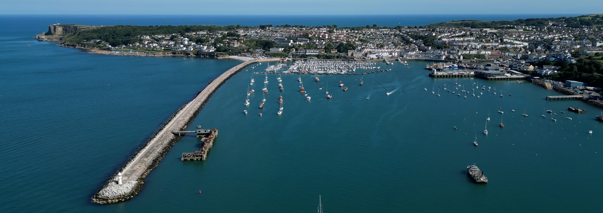 Aerial view of Brixham Harbour showing the long breakwater, marina filled with boats, moored yachts in the harbour, and the town rising along the South Devon coastline under clear blue skies.