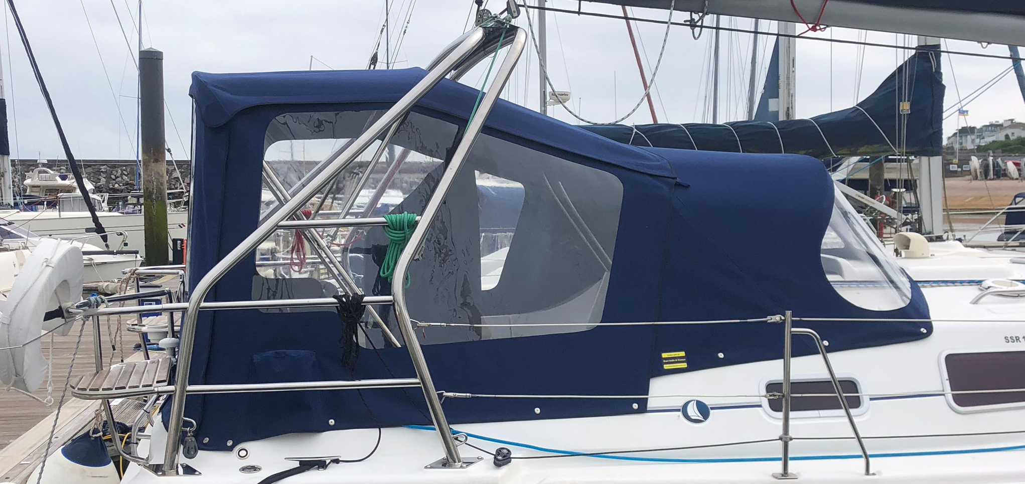 Custom navy-blue sprayhood and cockpit enclosure fitted to a sailing yacht, with clear windows providing protection from the elements while moored in a marina.