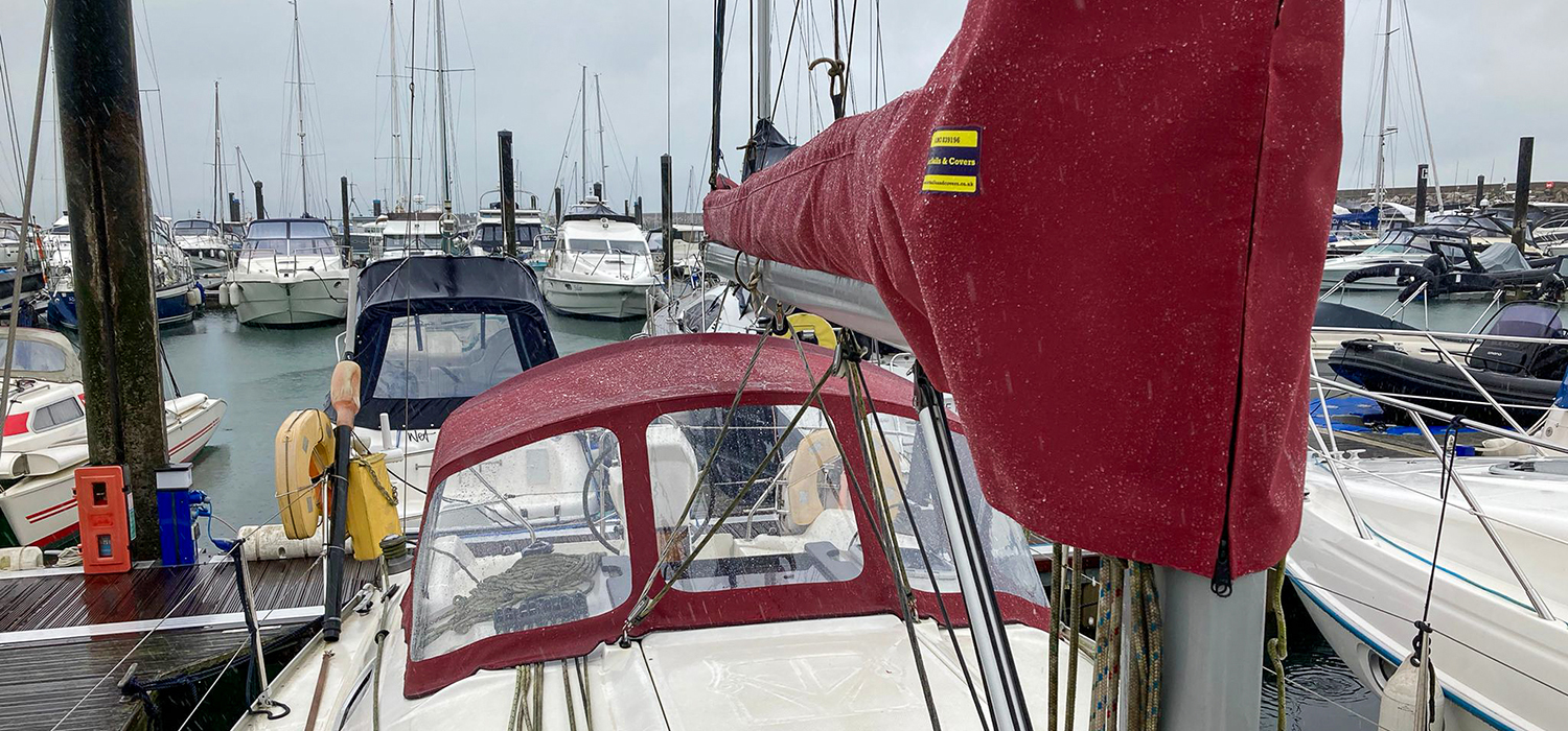 Sprayhood and sail cover Red Sprayhood and mainsail boom cover designed and made to measure by Dart Sails and Covers on a yacht in the marina, with raindrops beading on material due to its waterproof material